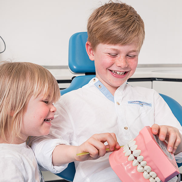 Kinder-in-der-Familienpraxis-Dr.-Gitt-quad Familienzahnarzt Dr. Gitt erklärt zwei Kindern das Zähneputzen anhand eines großen Zahnmodells.