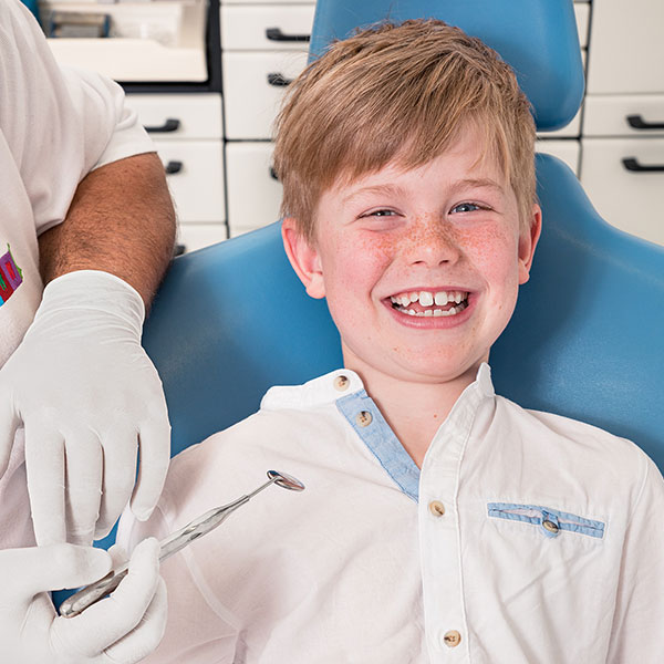 Zahnarzt-Dachau---Zahnarzt-Dr.-Gitt-behandelt-ein-Kind-1 Dr. Gitt, Zahnarzt Dachau, behandelt ein Kind auf einem blauen Zahnarztstuhl, hält zahnärztliche Instrumente in weißen Handschuhen. Der Junge lächelt nett.