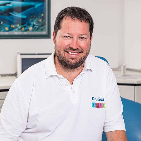 Zahnarzt-Dachau---Dr.-Michael-Gitt Dr.-Michael-Gitt, Zahnarzt Dachau, sympathisch lächelnd, mit Bart, trägt weißes Polohemd mit Aufschrift 'Dr. Gitt' und buntem Logo.