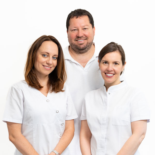 Team-Zahnarzt-Dachau-3 Das Zahnärzte-Team von Dr. Gitt - Zahnarzt Dachau. Team steht nebeneinander: Dr. Gitt in der Mitte, rechts und links sind zwei Zahnärztinnen zu sehen, alle mit Lächeln und weißem Kittel.