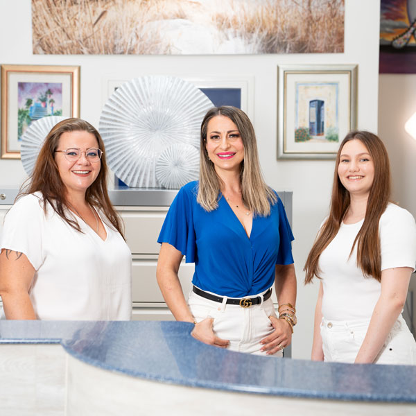 Zahnarztpraxis-Dachau---Empfang-2 Das Empfangsteam der Zahnarztpraxis Dachau (Saskia, Seher, Nudzejma) steht lächelnd hinter den Empfangstresen.