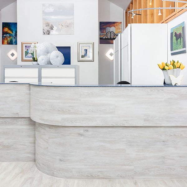 Zahnarzt-Dachau---Empfang Der Empfangstresen der Zahnarztpraxis Dachau, geschmackvoll eingerichtet mit moderner Holzverkleidung, Dekoration mit weißen Muscheln und gelben Tulpen, mehrere gerahmte Bilder an den Wänden.